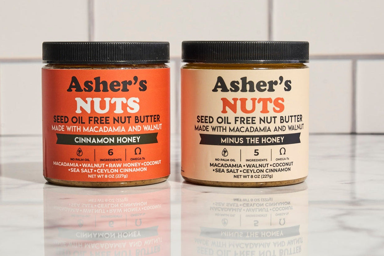 Two jars of Asher's Nuts nut butter on a marble surface with a tiled wall background.