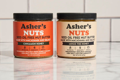 Two jars of Asher's Nuts nut butter on a marble surface with a tiled wall background.